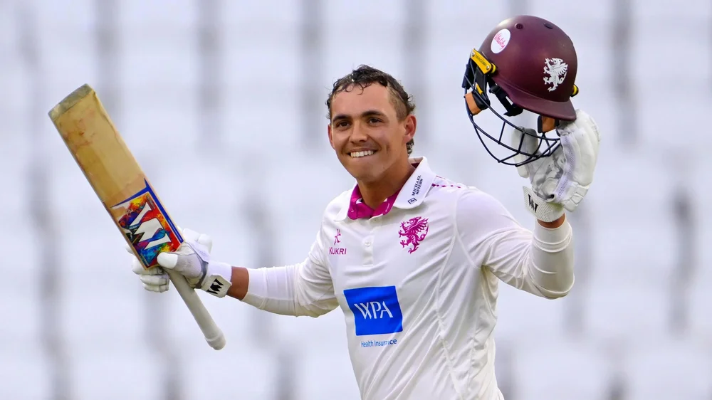 Josh Thomas holds his bat in the air in his right hand and his helmet in his left as he smiles after scoring a century against Yorkshire